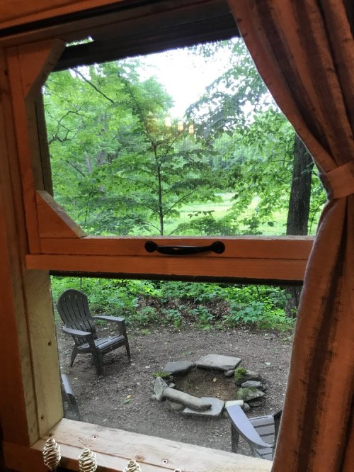 View from a window showing a wooded area with a fire pit and two chairs in a serene outdoor setting.