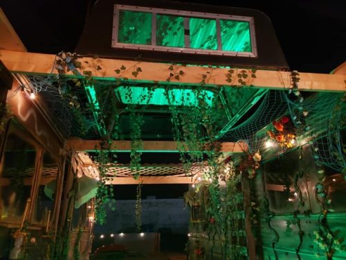 A cozy, illuminated space with green lighting, wooden beams, and hanging plants, creating a warm, inviting atmosphere.