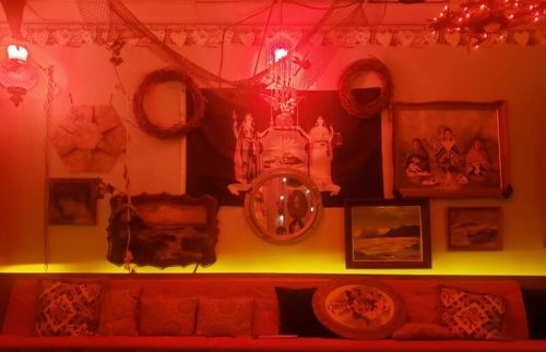 A cozy wall adorned with vintage art, mirrors, and decorative elements, illuminated by warm red lighting.