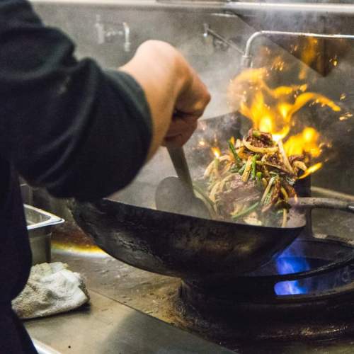 A chef stir-fries vegetables in a wok over a flame, creating a cloud of steam in a busy kitchen.