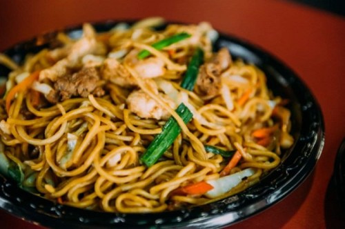 A close-up of a bowl of stir-fried noodles with vegetables and pieces of chicken on a red table.
