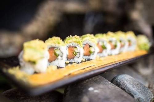 A close-up of a sushi roll on a black plate, featuring salmon, avocado, and a sprinkle of crunchy topping.