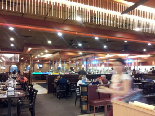 A busy restaurant interior with diners at tables, a buffet area, and a server carrying food. Warm lighting and decor.
