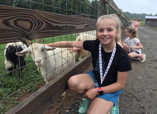 A girl smiles while petting a goat through a fence, with other children playing in the background on a farm.