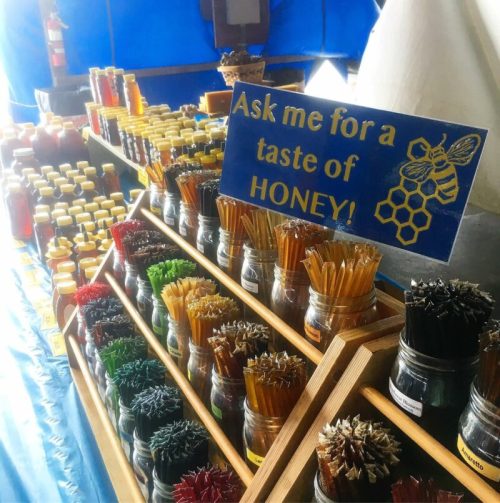 A display of honey jars and beeswax candles with a sign inviting customers to taste honey.