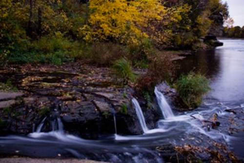 A serene river scene with gentle waterfalls, surrounded by autumn foliage and lush greenery.
