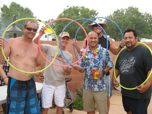 Five men at a backyard party hold colorful hula hoops, smiling and enjoying the festive atmosphere.