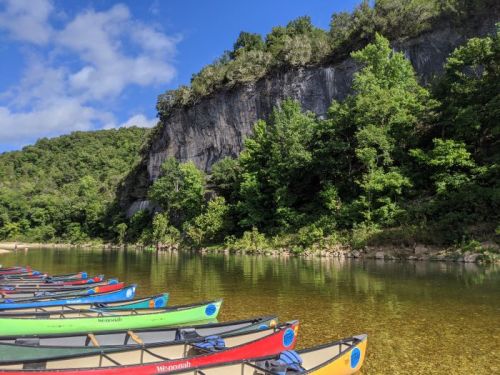 Colorful canoes lined up on a calm river, with lush greenery and a rocky cliff in the background under a blue sky.
