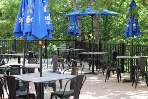 Outdoor dining area with black tables and chairs, blue umbrellas, surrounded by greenery.