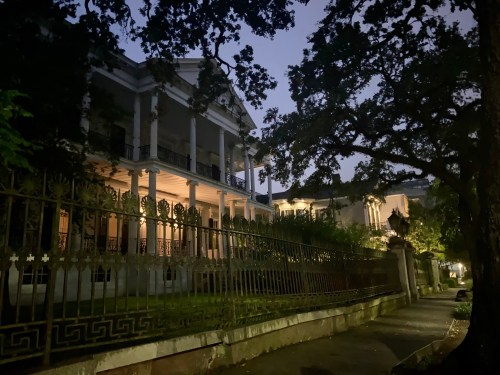 A historic mansion at dusk, surrounded by trees and a wrought iron fence, with soft lighting illuminating the facade.