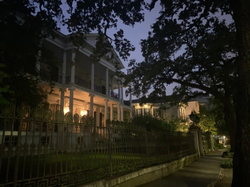 A historic mansion illuminated at night, surrounded by trees and a wrought iron fence, creating a mysterious atmosphere.