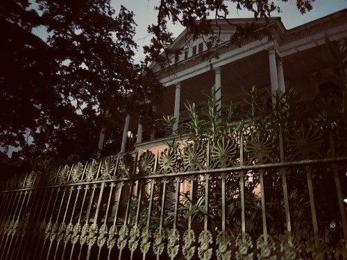 A dimly lit historic house behind an ornate iron fence, surrounded by trees at dusk.