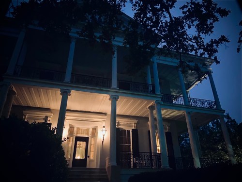 A large, historic house at night, illuminated by warm light, with tall columns and a dark, moody atmosphere.