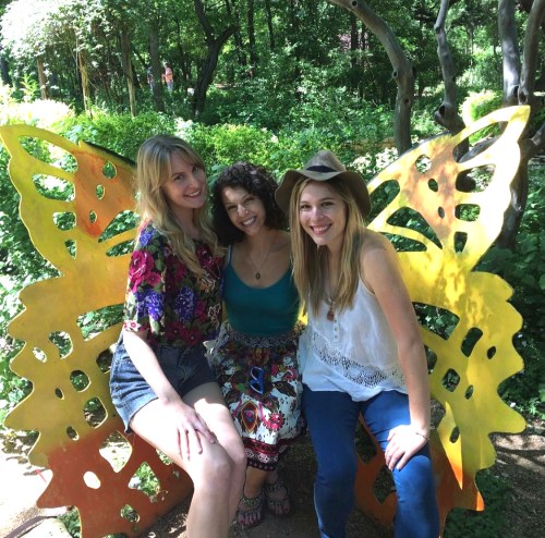 Three women sit on a colorful butterfly bench in a lush garden, smiling and enjoying a sunny day.