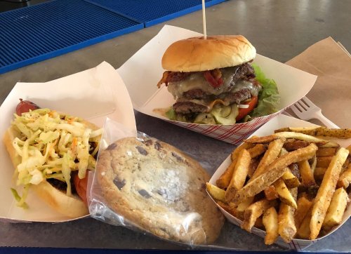 A burger with cheese and toppings, a hot dog with slaw, a cookie, and a side of seasoned fries on a table.