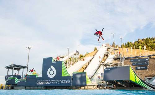 A skier performs a jump above a water ramp at Utah Olympic Park, with a large sign and spectators in the background.