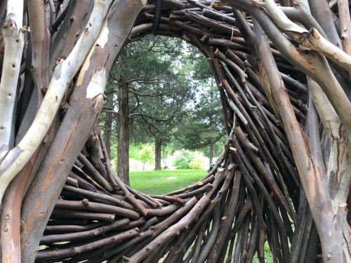 A circular structure made of intertwined branches, framed by trees and greenery in the background.