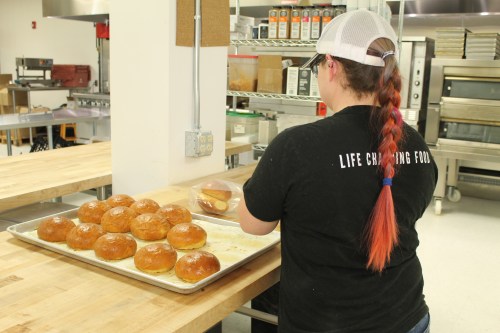A person with a braided hairstyle places freshly baked buns on a tray in a commercial kitchen.