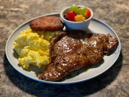 A plate with scrambled eggs, a steak, a hash brown patty, and a bowl of mixed fruit.