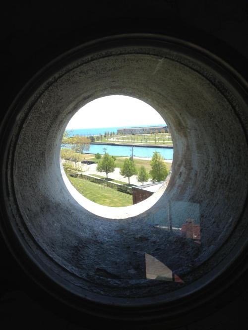 View through a circular opening, showcasing a park and water in the distance on a sunny day.