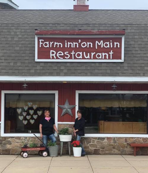 Two women stand outside the Farm inn'on Main Restaurant, surrounded by potted plants and a wagon.