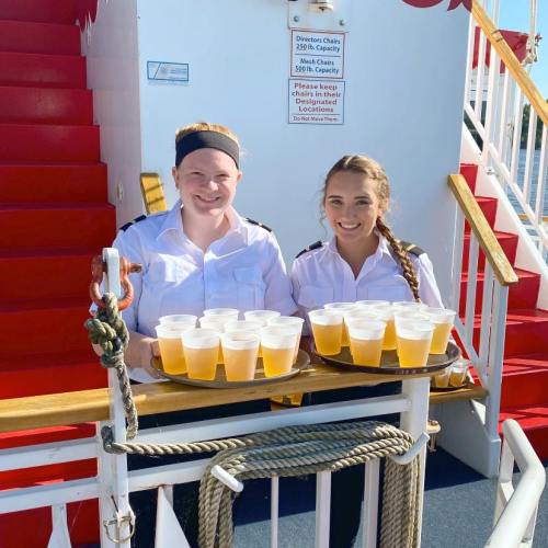 Two women in white shirts hold trays of drinks on a boat, with a red staircase and a sign in the background.