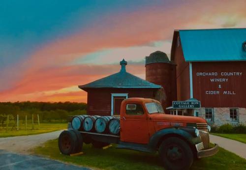 A vintage red truck parked in front of a rustic winery and cider mill at sunset, with vibrant skies and green fields.