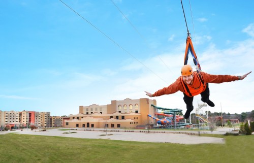 A person in an orange harness zips through the air on a zip line, with a resort and water park in the background.