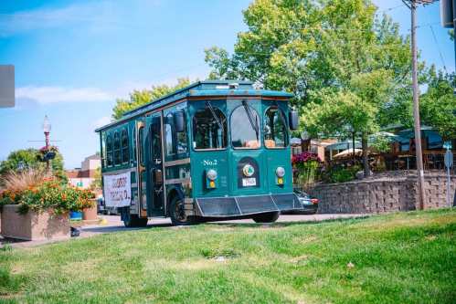 A green trolley bus labeled "No. 2" drives past a landscaped area with trees and flowers on a sunny day.