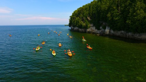 Aerial view of kayakers paddling in clear green waters near a rocky shoreline surrounded by lush trees.