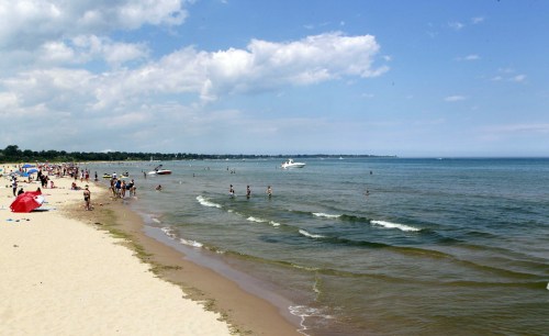 A sunny beach scene with people swimming, sunbathing, and boats on the water under a partly cloudy sky.