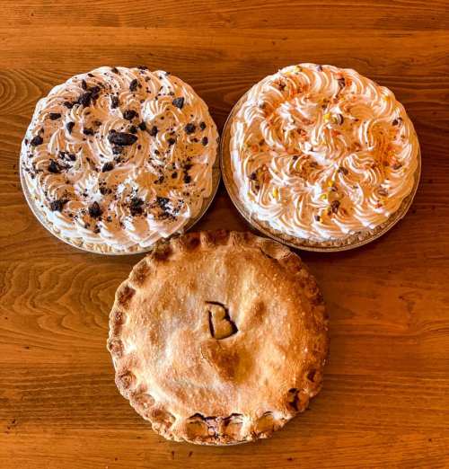 Three desserts on a wooden table: two cream-topped cakes and one fruit pie with a decorative crust.