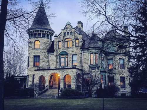 A large, ornate stone mansion with a turret, illuminated windows, and a twilight sky in the background.