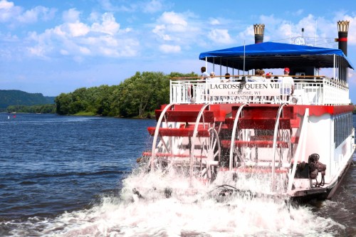 A paddleboat named Lacrosse Queen VIII navigates a river, with lush greenery and blue skies in the background.