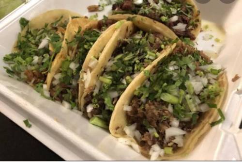 Five tacos filled with chopped meat, topped with onions and fresh cilantro, served in a takeout container.