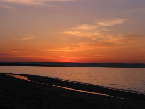 A serene sunset over a calm lake, with soft orange and pink hues reflecting on the water and a sandy shore.