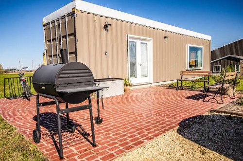 A modern shipping container home with a patio, grill, and seating area, set in a sunny outdoor space.