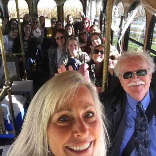 A group of smiling people on a bus, with a woman taking a selfie in the foreground and a driver in a suit.
