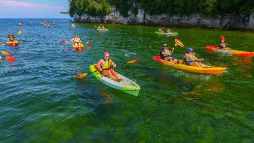 A group of people kayaking in clear green water, surrounded by rocky cliffs and lush greenery on a sunny day.