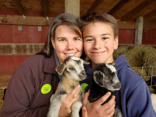 A woman and a boy smile while holding two baby goats in a barn setting.