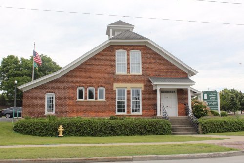 Historic brick building with a gabled roof, large windows, and a front staircase, surrounded by greenery and a flagpole.