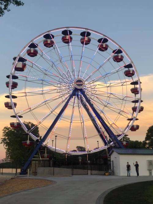 A brightly lit Ferris wheel at sunset, with colorful gondolas against a scenic sky. Two people walk nearby.