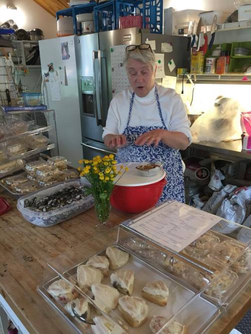 A woman in an apron prepares baked goods in a kitchen, surrounded by trays of treats and a vase of flowers.