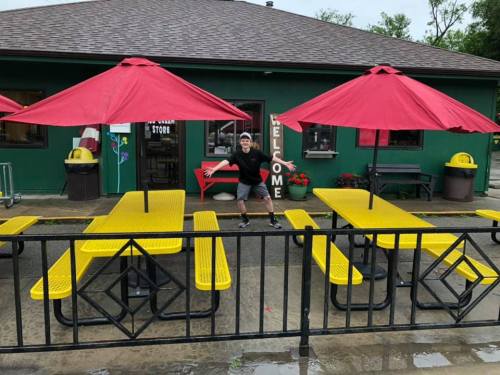 A young person poses playfully in front of yellow picnic tables with red umbrellas outside a green building.