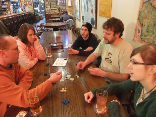 A group of five people playing a board game at a wooden table in a cozy bar, with drinks in front of them.