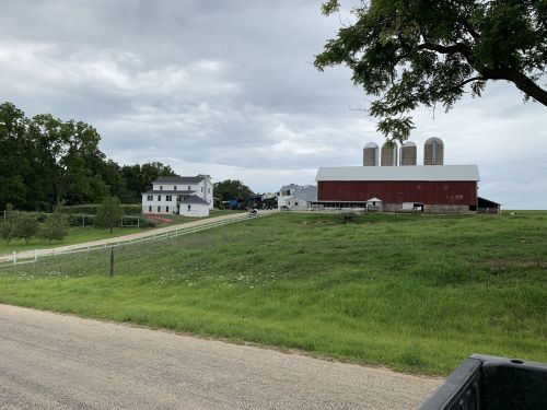 A rural farm scene featuring a red barn, silos, and white farmhouse under a cloudy sky, surrounded by green fields.