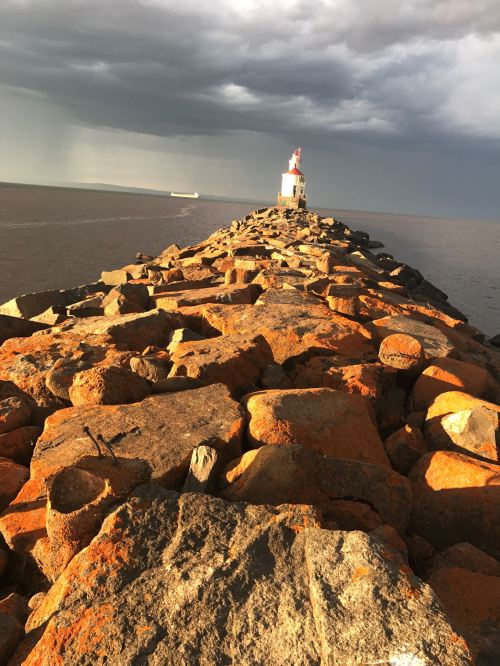 A rocky jetty leads to a small lighthouse under a cloudy sky, with a distant ship on the horizon.