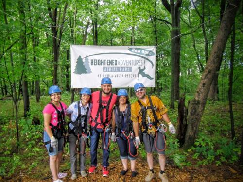 A group of five people in harnesses and helmets stand in front of a "Heightened Adventures" sign in a forested area.