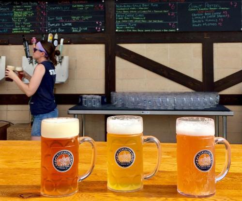 Three frosty beer mugs in front of a bar, with a bartender in the background and a menu board above.