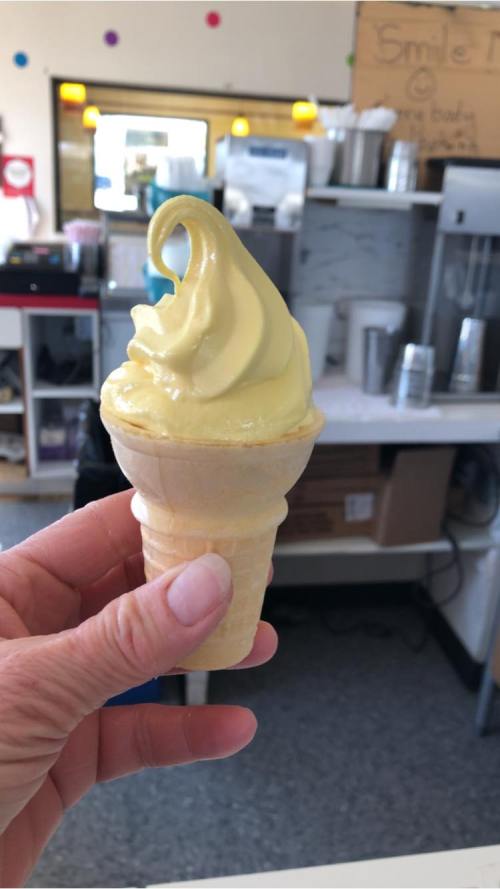 A hand holding a soft-serve ice cream cone with a yellow swirl, set against a café background.
